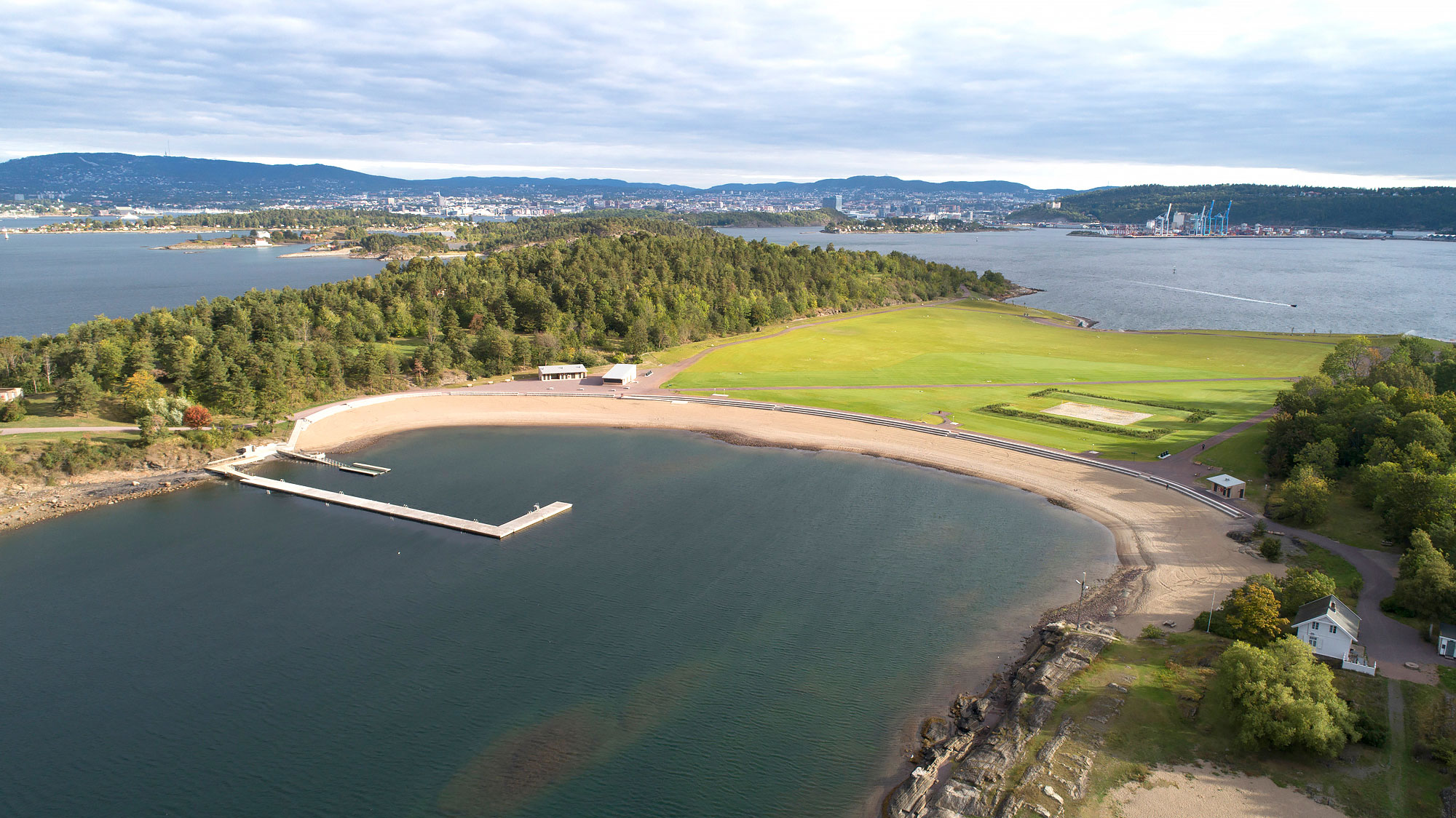 Dronebilde av Nye Langøyene viser en u-formet tilrettelagt badestrand i forkant av en gjennomgående gresslette, omgitt av løvskog. Utformet av landskapsarkitekter og et tverrfaglig teamFoto: Asplan Viak/Jostein Thorvaldsen