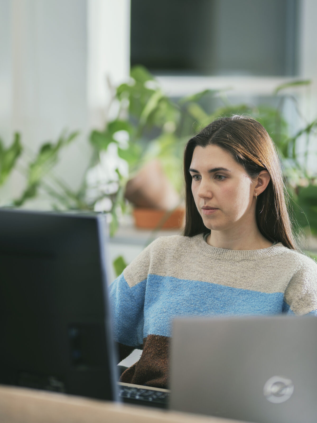 en person som sitter i et kontorlandskap foran en pc og med grønne planter bak seg. Bærekraftsrapportering.
