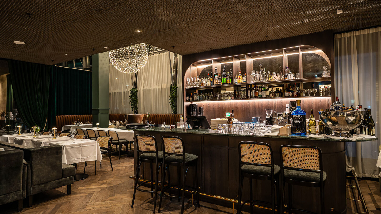 Fra restauranten i Sixa mot barområdet med sin belysning. Flere bord og stoler og en stor rund lampe som lyser.