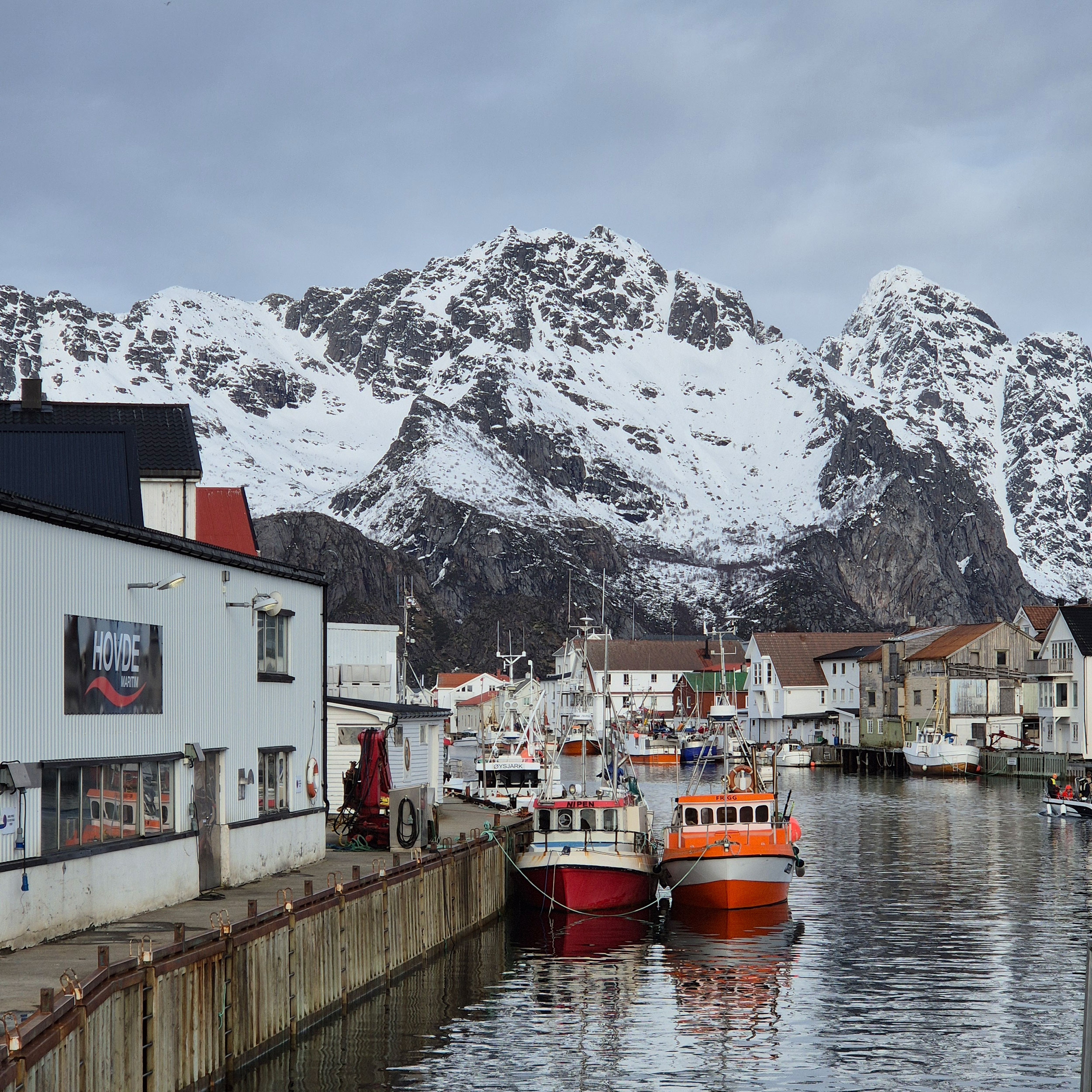 Trehusbegyggelse i lofoten ved båthavn med fjell i bakgrunnen.