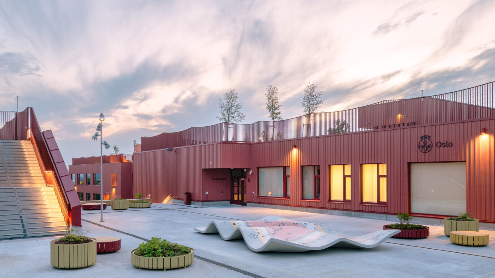 Afternoon view of a school with red timber cladding and a flat blue-green roof planted with trees and shrubs. The schoolyard features planters and a play sculpture resembling a flying carpet.