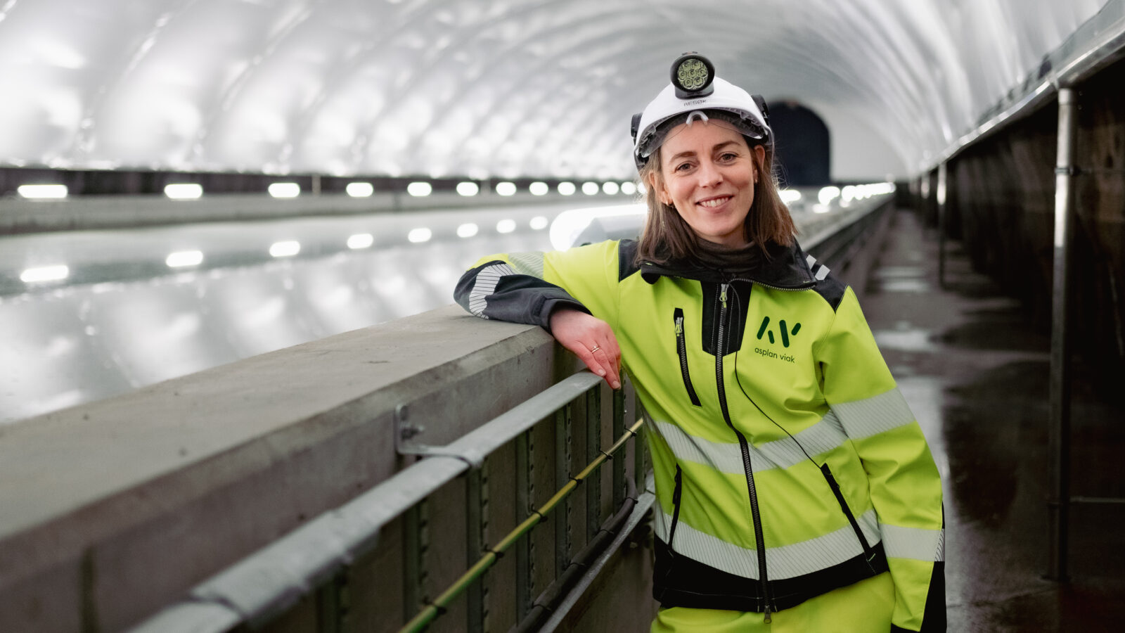 A young female civil engineer in high-visibility workwear and a safety helmet with a headlamp, standing beside a concrete structure inside a water supply facility.