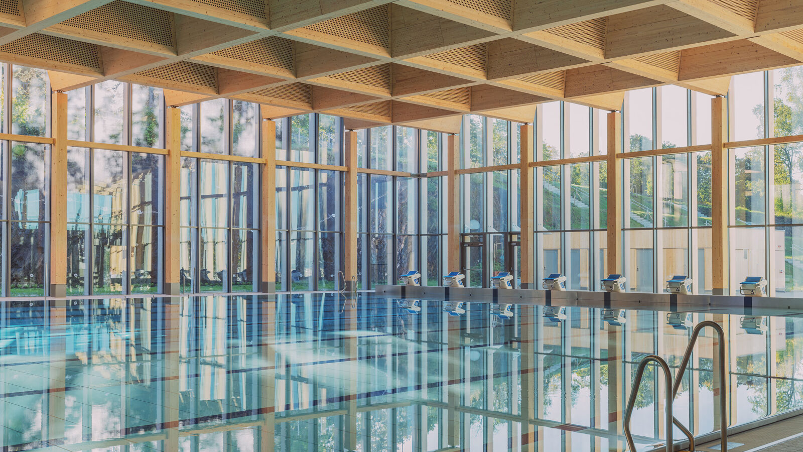 The corner of a large indoor swimming pool with starting blocks and glazed façades along both walls. The mass timber ceiling features large, square recesses that conceal ventilation systems.