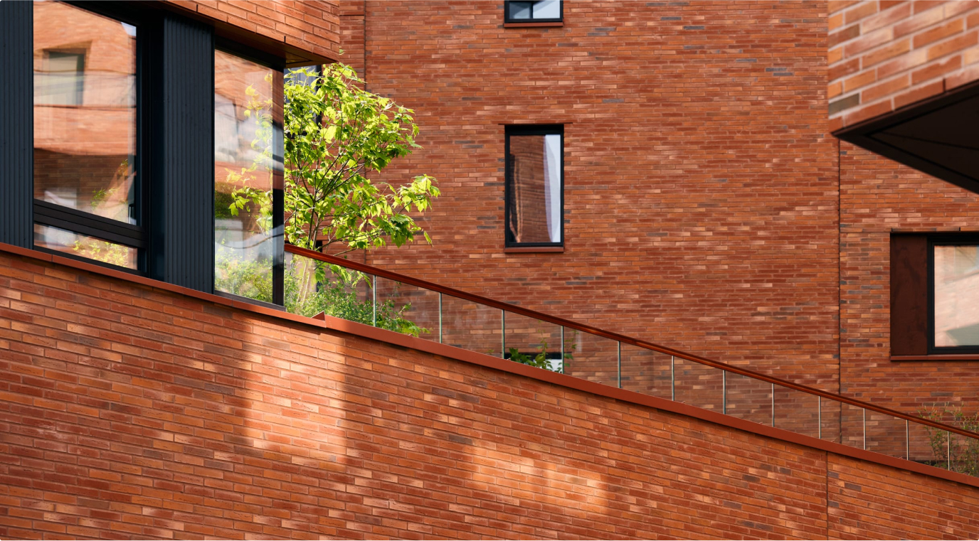 Detail of a red brick building with an upper-floor terrace enclosed by a glass balustrade, with trees and planting on the terrace.
