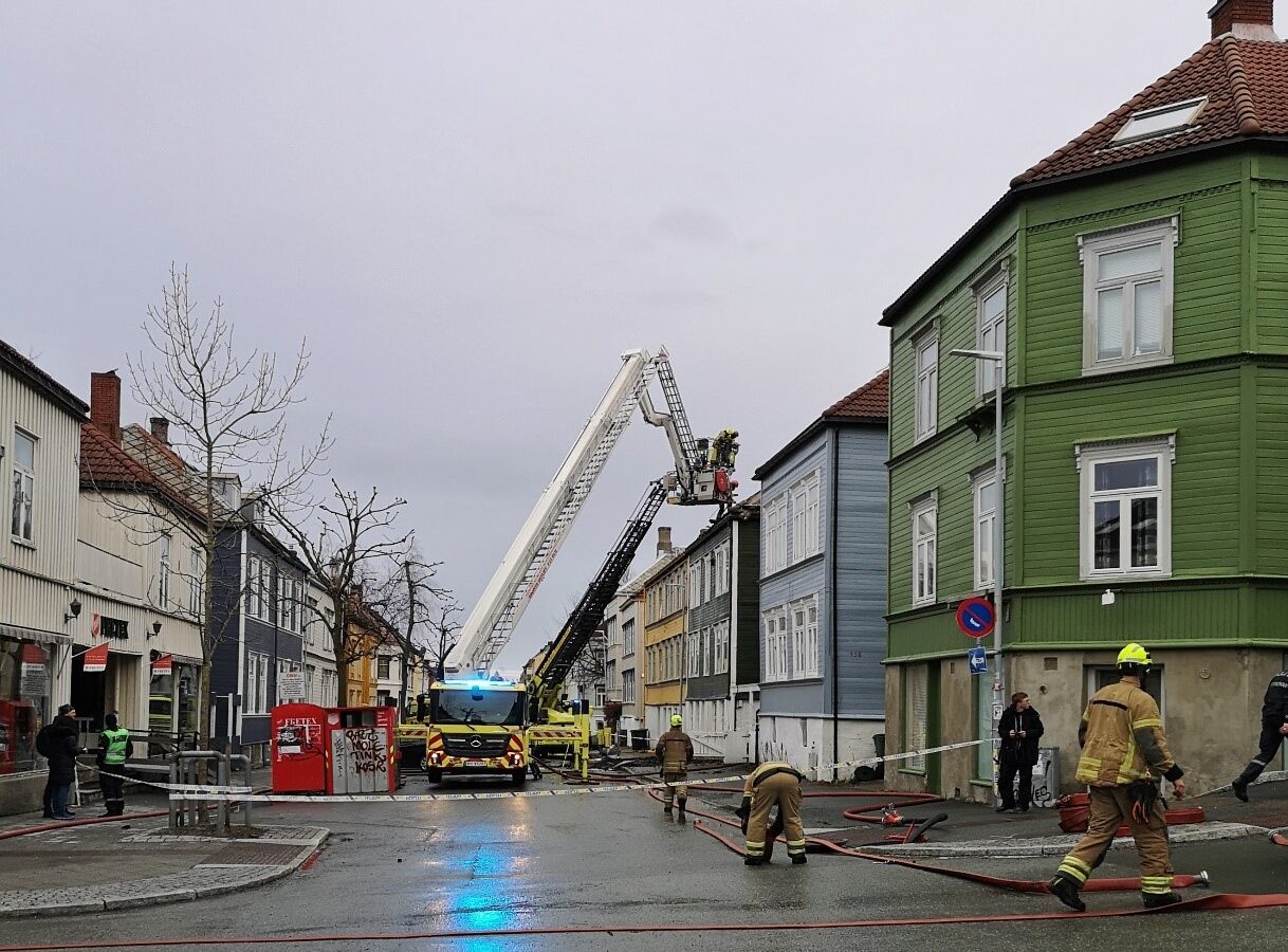 A street lined with wooden houses is cordoned off with police tape. Two fire engines with aerial ladders and firefighters tackling a blaze in one of the houses are visible beyond the cordon.