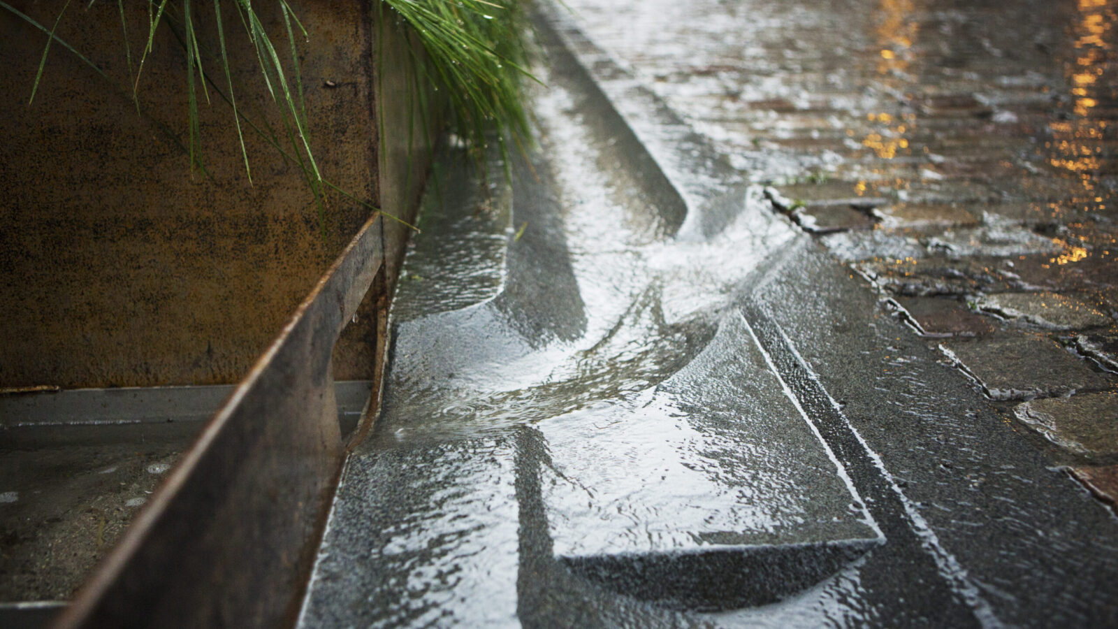 Close-up of a cobblestone street edge, where carved channels direct rainwater into a storm drain.