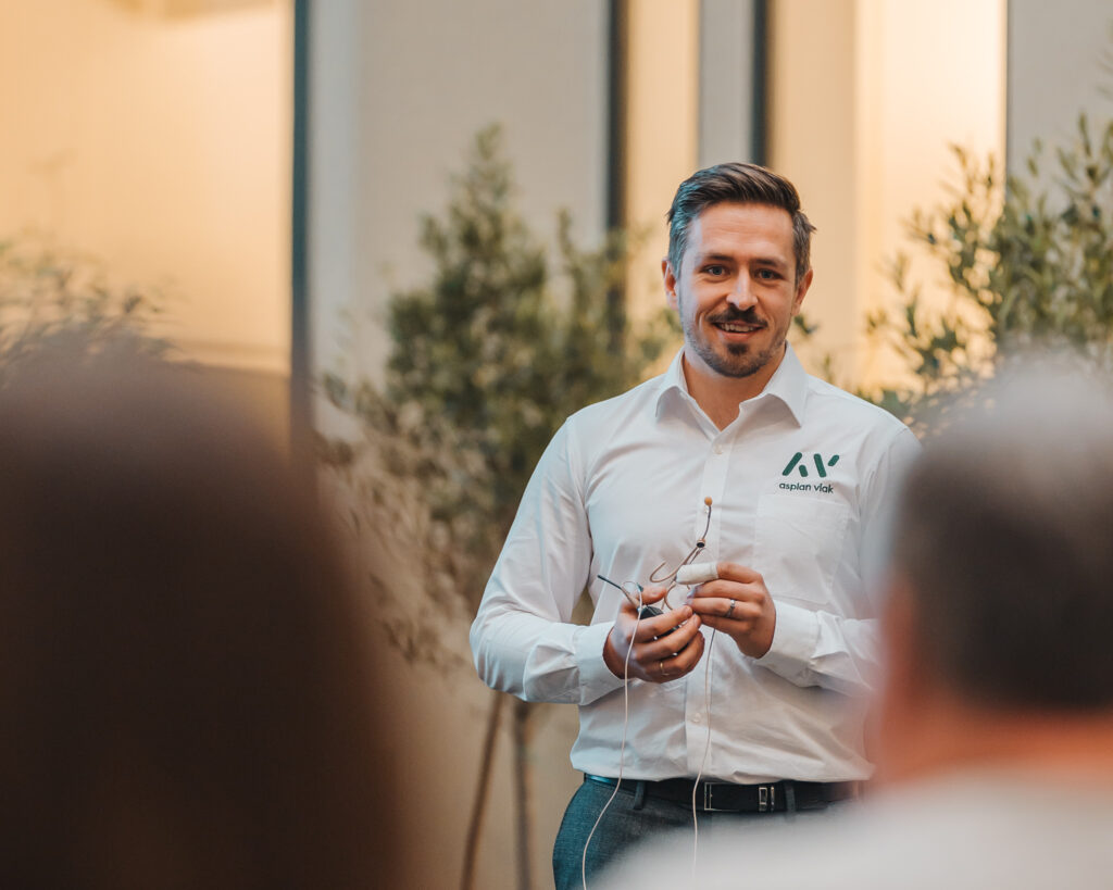 A smiling man with the AV logo on his shirt holds a microphone and speaks to a group of people with their backs to the camera.